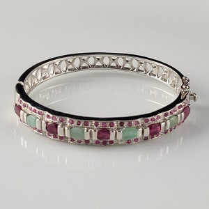 May include: A silver bangle bracelet with a delicate openwork design and a row of alternating green and red gemstones.
