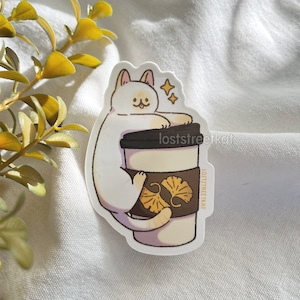 May include: White cat sticker with a brown and white coffee cup with a ginkgo leaf design. The text "loststreetkat" is on the sticker.