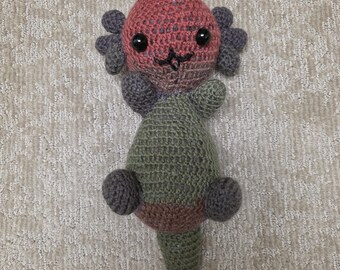 May include: A handmade crochet axolotl plushie in shades of pink, gray, and green. The axolotl has a round head with black safety eyes, gray gills, and a stitched mouth. The body is green with a brown stripe.