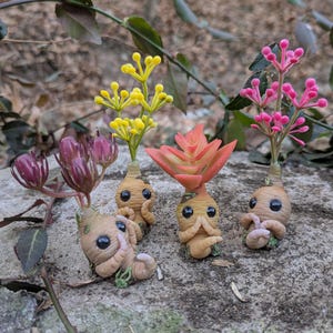 May include: A collection of handmade clay figurines resembling Mandrakes, each with a unique floral head. The figurines are tan with expressive eyes and are posed on a stone surface. The flowers are yellow, pink, and orange.