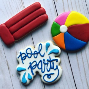 May include: Three decorated sugar cookies. One cookie is red and shaped like a pool float. Another cookie is blue, yellow, orange, green, and pink and shaped like a beach ball. The third cookie is white with blue lettering that says "pool party" and blue water splashes.