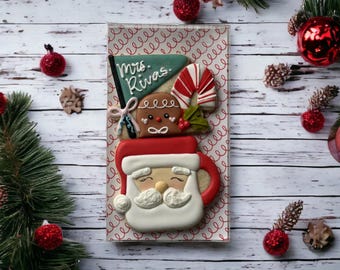 May include: A festive Christmas cookie arrangement featuring a Santa mug, gingerbread man, candy cane, and a banner that reads "Mrs. Rivas." The cookies are decorated with red, white, and brown icing, and are presented in a clear box. The background includes Christmas ornaments and pine needles.