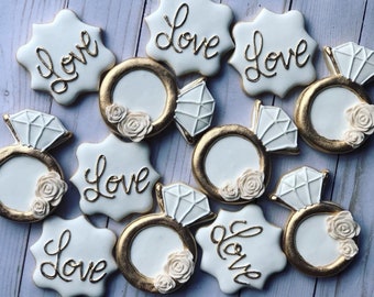 Puede incluir: Un conjunto de diez galletas de azúcar blancas y doradas decoradas con la palabra "Love" y un anillo con un diamante. Las galletas son perfectas para una boda o una fiesta de compromiso.