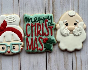 May include: Three Christmas-themed cookies. One is a Mrs. Claus face with glasses and a red bow. The middle cookie says "Merry Christmas" in red letters on a green background. The last cookie is Santa's face.