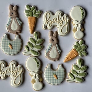 May include: Decorated sugar cookies in a baby shower theme. The cookies include bunny, carrot, bib, rattle, and leaf shapes. The word "baby" is written in cursive. The cookies are decorated with white, tan, green, and orange icing.