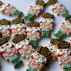 May include: A collection of decorated sugar cookies shaped like nutcracker soldiers. The cookies are decorated with pink, green, gold, and white icing.
