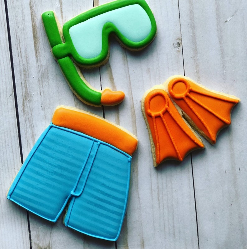 May include: A set of four sugar cookies decorated with icing to look like summer swimming gear. The cookies are shaped like a pair of swim trunks, a snorkel and goggles, and a pair of flippers. The swim trunks are blue with orange trim, the snorkel and goggles are green, and the flippers are orange.