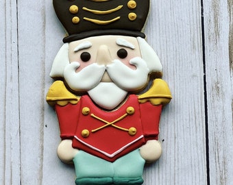 May include: A decorated gingerbread cookie shaped like a nutcracker. The cookie is decorated with red, white, green, and yellow icing. The nutcracker has a black hat with gold trim, a white beard, and a red and white uniform.