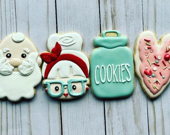 May include: Four decorated sugar cookies. One cookie is a heart shape with pink icing and sprinkles. Another cookie is a jar with blue icing and the word "COOKIES" in white icing. The third cookie is a cartoon of a woman with red hair and glasses. The fourth cookie is a cartoon of a man with a white beard and a red hat.