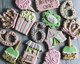 May include: A set of 11 pink, green, and gold decorated sugar cookies with various circus themes, including a popcorn cart, a tent, a crown, a cotton candy, and the number six. The cookies are decorated with sprinkles, gold accents, and the text "ADMIT ONE" and "Layla".