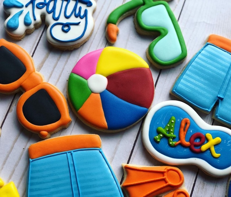 May include: A set of nine colorful sugar cookies decorated with summer themes. The cookies include a beach ball, sunglasses, a pool float, and swim trunks. The cookies are decorated with blue, green, orange, red, and yellow icing.