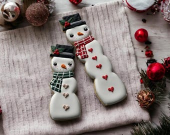 May include: Two decorated sugar cookies shaped like snowmen. The larger snowman is white with red hearts and a red and white scarf. The smaller snowman is white with green and white plaid scarf and red hearts. Both snowmen have black hats and black eyes.