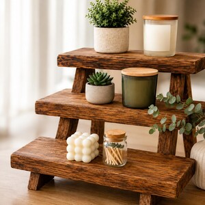 Puede incluir: Un expositor de madera de tres niveles con un acabado texturizado en marr&oacute;n oscuro. Los estantes contienen una peque&ntilde;a planta en maceta, una vela con tapa de madera, una suculenta en maceta gris, una vela verde, una vela de burbujas y un frasco.