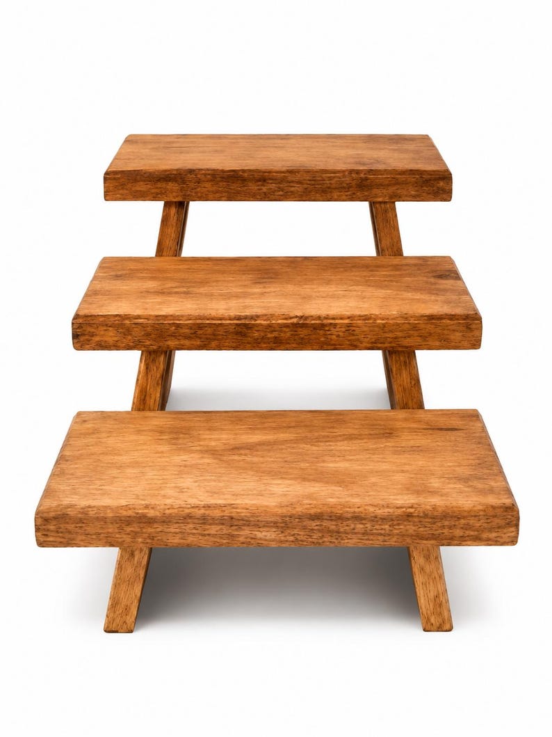 Puede incluir: Un expositor de madera de tres niveles con plataformas rectangulares y un acabado de veta de madera marr&oacute;n medio. Cada nivel est&aacute; sostenido por cuatro patas inclinadas, creando una estructura escalonada que aumenta en altura hacia atr&aacute;s.