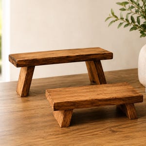 Puede incluir: Dos elevadores rectangulares de madera con un acabado marr&oacute;n natural y veta de madera visible. Cada elevador cuenta con una superficie superior plana sostenida por dos patas en &aacute;ngulo, proporcionando una base estable para exhibici&oacute;n.