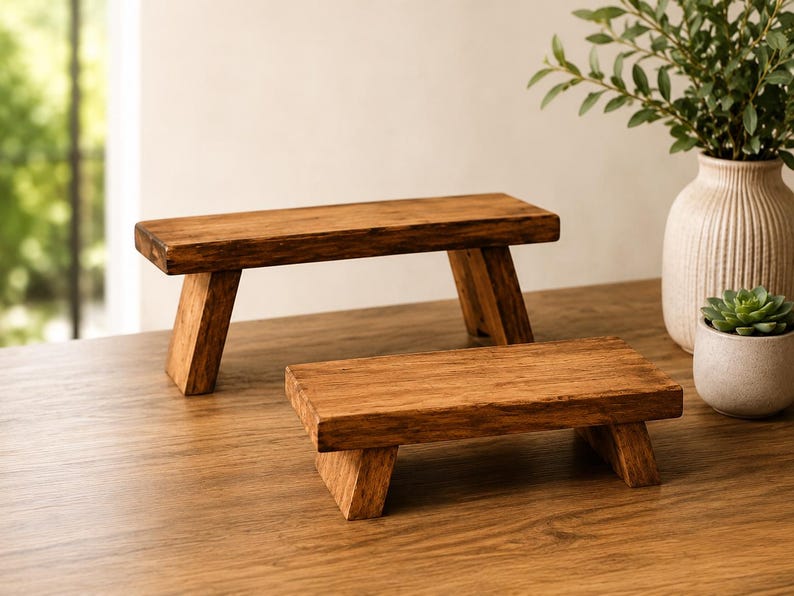 Puede incluir: Dos elevadores rectangulares de madera con un acabado marr&oacute;n natural y veta de madera visible. Cada elevador cuenta con una superficie superior plana sostenida por dos patas en &aacute;ngulo, proporcionando una base estable para exhibici&oacute;n.