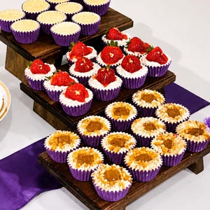 Puede incluir: Un expositor de madera de tres niveles con varios pastelitos en moldes de papel morado. El nivel superior tiene pastelitos de color crema, el nivel medio tiene pastelitos con fresas frescas y el nivel inferior tiene pastelitos con un centro de color caramelo.