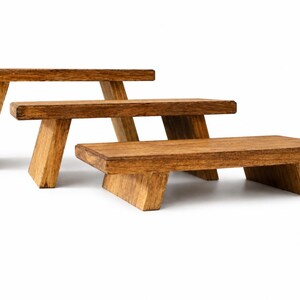 Puede incluir: Tres expositores rectangulares de madera con un acabado marr&oacute;n medio y veta visible. Cada expositor cuenta con una superficie plana sostenida por dos patas inclinadas, dispuestos en una formaci&oacute;n escalonada de mayor a menor.