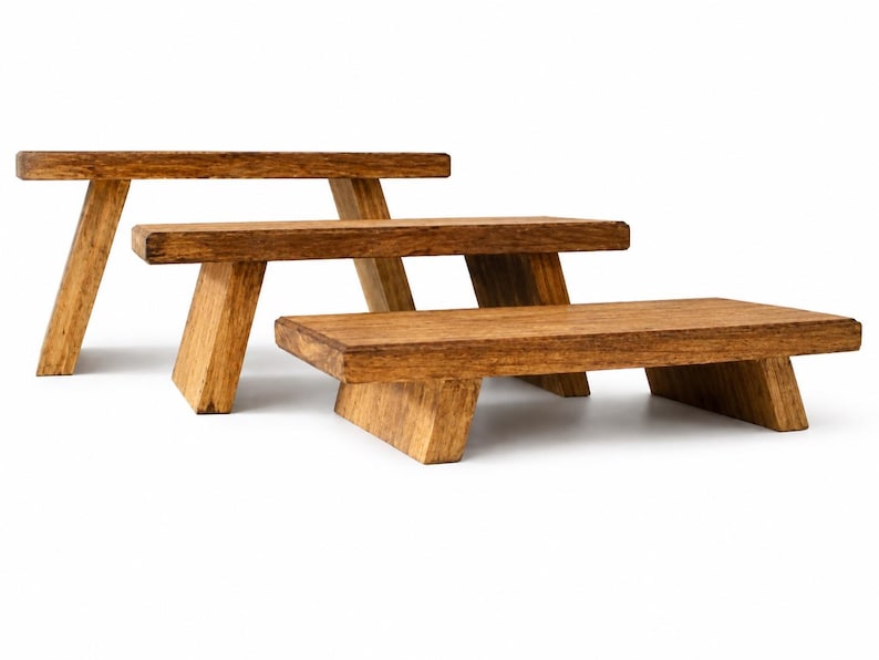 Puede incluir: Tres expositores rectangulares de madera con un acabado marr&oacute;n medio y veta visible. Cada expositor cuenta con una superficie plana sostenida por dos patas inclinadas, dispuestos en una formaci&oacute;n escalonada de mayor a menor.
