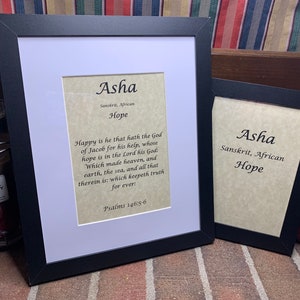 May include: Two framed prints with the name "Asha" and the word "Hope." The larger frame has a white mat and a black frame. The text includes a Sanskrit and African origin, along with a Bible verse. The smaller frame is black.