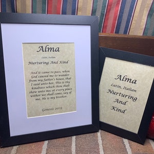 May include: Two framed prints with the name "Alma" in a black frame. The prints include the words "Latin, Italian, Nurturing And Kind" and a Bible verse from Genesis 20:13. The frames are on a brick surface.