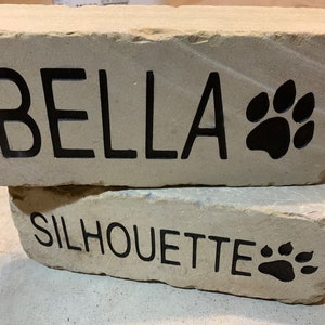 May include: Two stacked beige stone bricks with black lettering. The top brick reads "BELLA" with a black paw print. The bottom brick reads "SILHOUETTE" with a black paw print.
