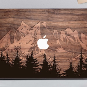 Könnte beinhalten: Eine Laptop-Hülle aus Holz mit einem Bergketten-Design und einer Waldsilhouette. Das Apple-Logo ist weiß. Die Hülle hat eine natürliche Holzmaserung und eine braune Farbe.