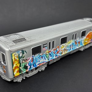 May include: A silver toy subway car model featuring colorful graffiti art along the side. The car is marked with the number 1668 and a small American flag decal. It has a ribbed roof, rectangular windows, and detailed undercarriage components.