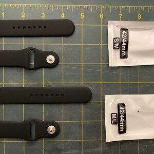 May include: Two sets of black silicone watch bands for 42/44mm devices, displayed on a green cutting mat with a yellow grid. One set is labeled S/M and the other M/L. Each set includes a strap with pin holes and a strap with a metal pin closure.
