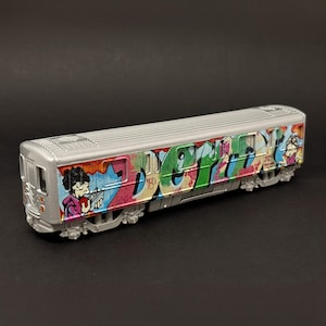 May include: A silver model subway car featuring colorful graffiti art along its side. The design includes stylized lettering in shades of green, pink, and blue, alongside a cartoon character illustration wearing a purple hoodie and holding a red object.