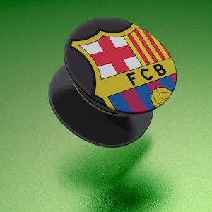 May include: A black phone grip with the FC Barcelona logo. The logo features a shield design with a red cross, yellow stripes, and blue and red stripes. The letters "FCB" are also visible.