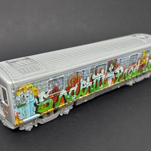 Puede incluir: Un modelo de vagón de metro de juguete de color plateado con arte de grafiti detallado en el lateral. La ilustración incluye letras verdes vibrantes, un personaje de dibujos animados y un patrón de pared de ladrillo, techo acanalado y ruedas.