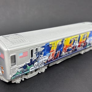 Puede incluir: Un modelo de vagón de metro plateado con coloridas ilustraciones estilo grafiti en el lateral. El diseño incluye patrones azules, amarillos y rojos con pequeñas calcomanías de banderas estadounidenses cerca del frente y texto integrado en el diseño.
