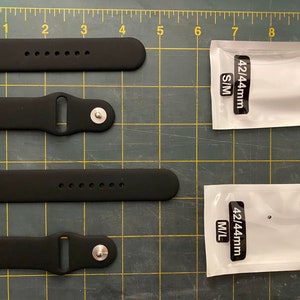 Puede incluir: Cuatro correas de reloj de silicona negra de diferentes tama&ntilde;os, dos paquetes de correas de reloj etiquetados "42/44mm S/M" y "42/44mm M/L" en una cuadr&iacute;cula negra y blanca.