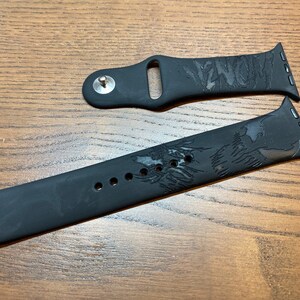 May include: A black silicone watch band featuring an engraved Venom character graphic and the word Venom. The two-piece strap includes a metal pin closure and attachment lugs designed for a smartwatch, showcasing a matte finish and detailed etching.