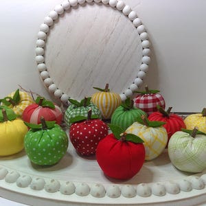 May include: A collection of colorful fabric apples with green stems and leaves, arranged on a white, decorative tray. The apples feature various patterns, including polka dots, stripes, and gingham. A white beaded circle is in the background.