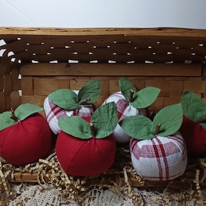 May include: A collection of six fabric apples featuring solid red and red-and-white plaid patterns. Each apple is topped with two green fabric leaves and a small brown twig stem. The apples are arranged inside a woven wooden basket.