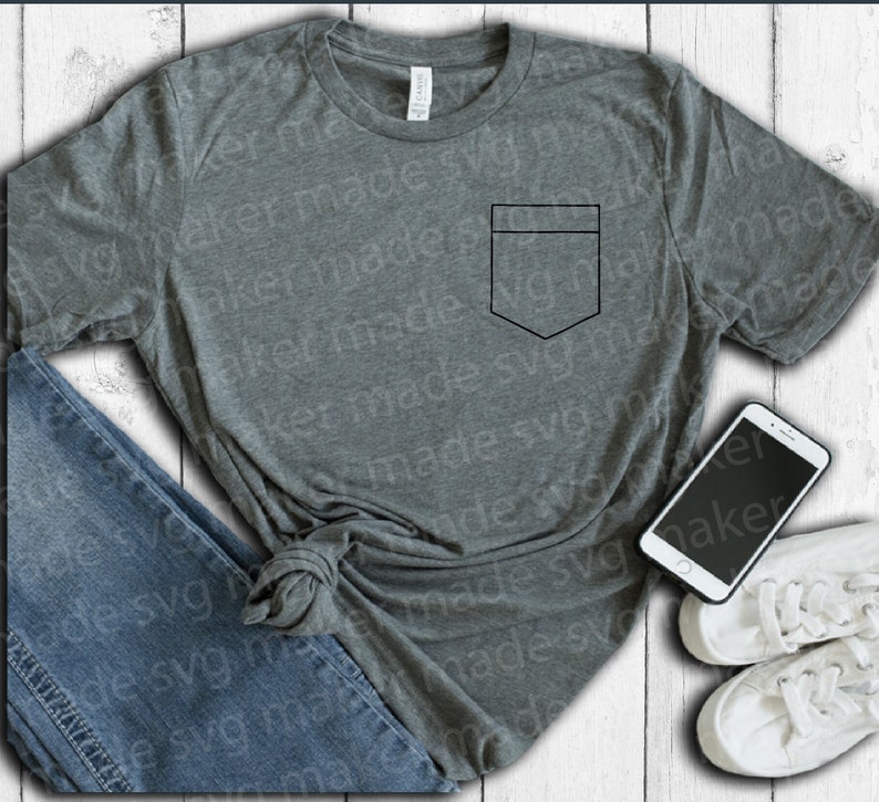 May include: A gray t-shirt with a black pocket design, paired with blue jeans, white sneakers, and a smartphone. The shirt is laid on a white wooden surface.