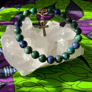 May include: A beaded bracelet featuring green and blue spherical stones. A gold-colored ankh charm hangs from the bracelet. The bracelet is displayed on a white crystal against a colorful patterned fabric.