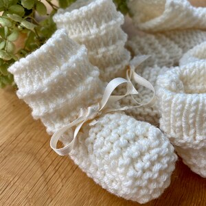 May include: A pair of white knitted baby booties with a satin ribbon tied around the ankle.