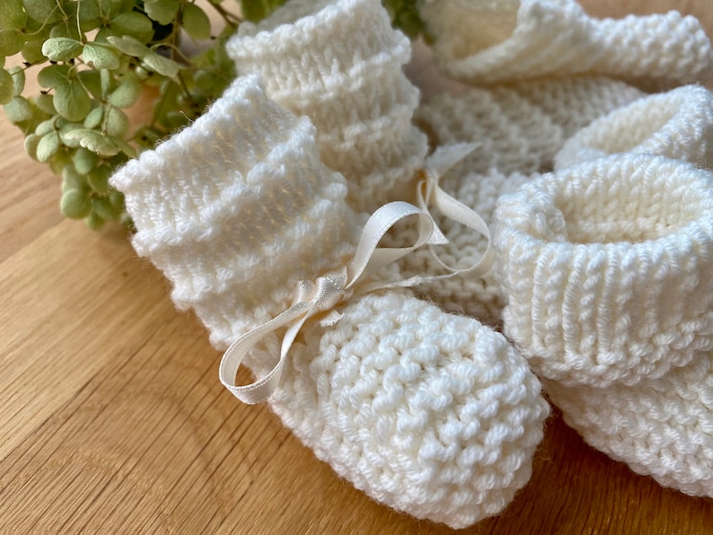 May include: A pair of white knitted baby booties with a satin ribbon tied around the ankle.