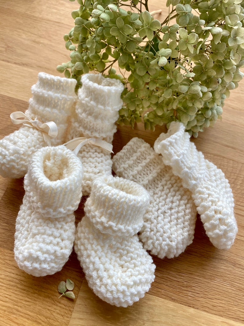 May include: A set of four white knitted baby booties with a ribbed texture. The booties are arranged on a wooden surface with a bouquet of white flowers in the background.