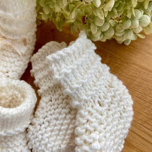 May include: A pair of white knitted baby booties with a delicate stitch pattern. The booties are soft and cosy, perfect for keeping little feet warm.