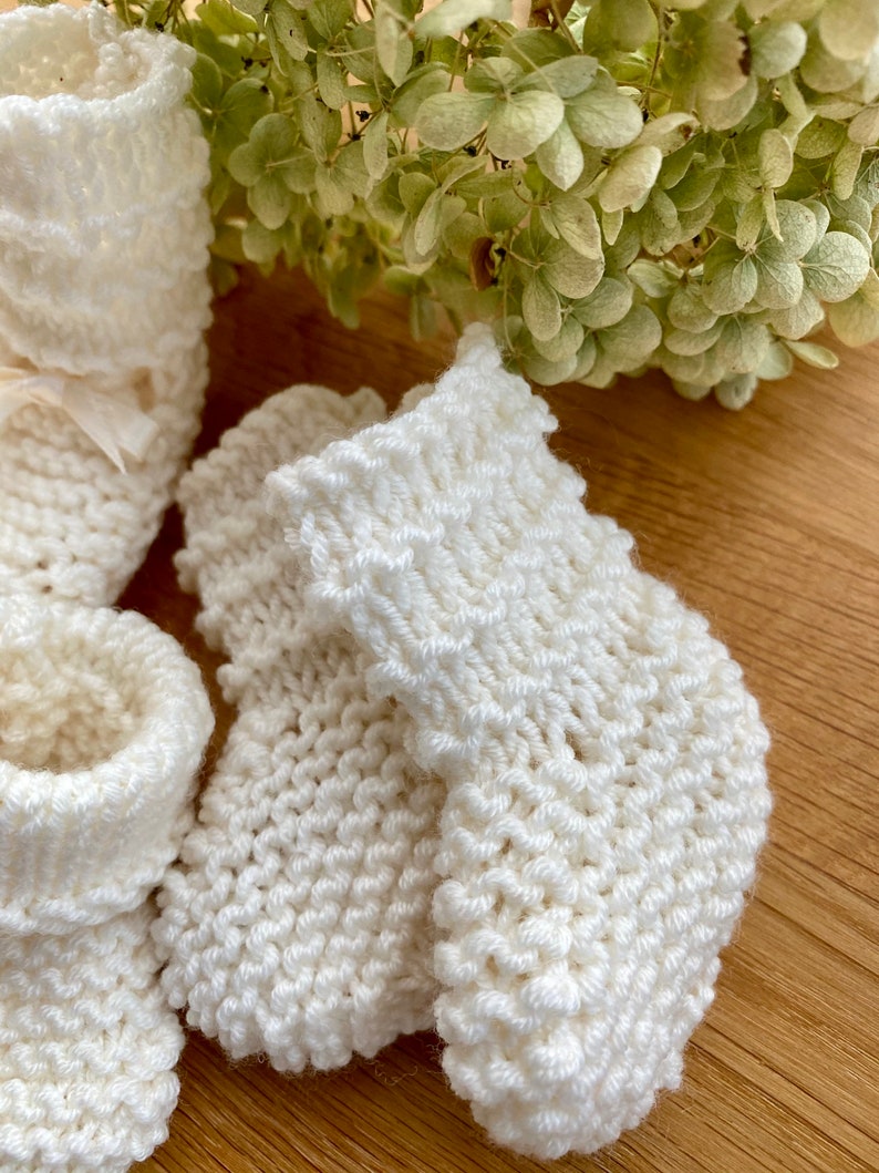 May include: A pair of white knitted baby booties with a delicate stitch pattern. The booties are soft and cosy, perfect for keeping little feet warm.
