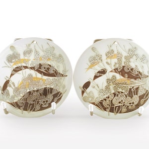 May include: Two round, off-white decorative plates with a brown and gold leaf design. The plates are held by white metal stands. The design features stylized leaves and floral elements, creating a nature-inspired aesthetic.