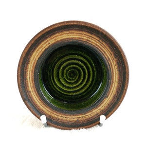 May include: A small, round ceramic bowl with a spiral design in the centre. The bowl features concentric rings of brown, tan, and green. The interior is a deep green, and the exterior has a textured, earthy appearance.