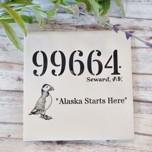 May include: White ceramic tile with black text and a black and white illustration of a puffin. The text reads "99664 Seward, AK "Alaska Starts Here""