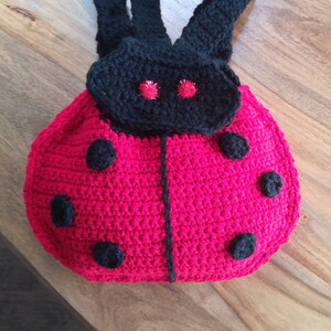 May include: A ladybug-shaped crochet backpack featuring a vibrant red body with black circular spots, a black head section with two small red metallic eyes, and two black crochet shoulder straps attached to the top of the bag.