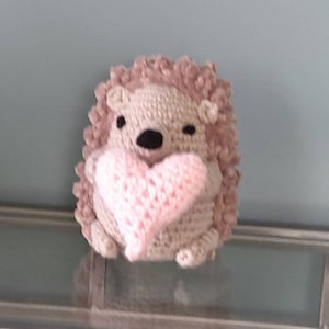 May include: A handmade crochet hedgehog holding a pink heart. The hedgehog has a light brown body, a darker brown back, and a black nose and eyes. The heart is a soft pink color. The toy is likely a decorative item or a gift.