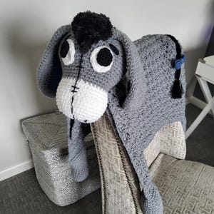 May include: A crocheted Eeyore blanket in shades of gray, white, and black, draped over a patterned chair. The blanket features Eeyore's head with large eyes and a fluffy black mane. The tail has a blue bow and a black tassel.
